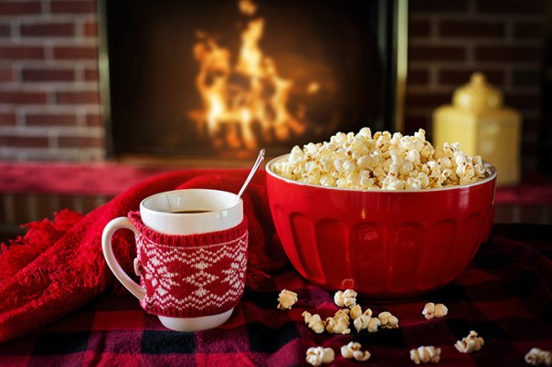 Popcorn in a red bowl and a mug of a warm beverage on the table with a fireplace in the background.