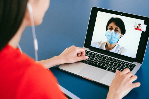 two women on a video conference. The view is over one woman's shoulder and you can see the other woman on the computer screen. The woman on the computer screen is a doctor wearing a surgical mask and doctor's white coat.