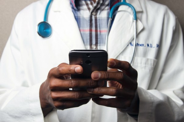 A man with only his Torso visible. He is wearing a doctor’s coat and stethoscope around his neck. He is holding a cell phone in his hands.