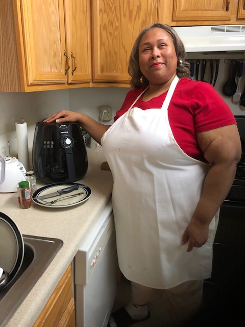 Empish is smiling and standing in kitchen with hand on her air fryer. She is wearing a white kitchen apron and the air fryer is on the counter with plate and seasonings.