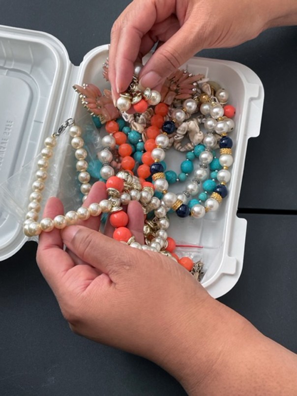 A small plastic rectangular box with an assortment of jewerly. It has several pieces of costume jewerly in bright colors. Hands are in the box selecting a necklace.