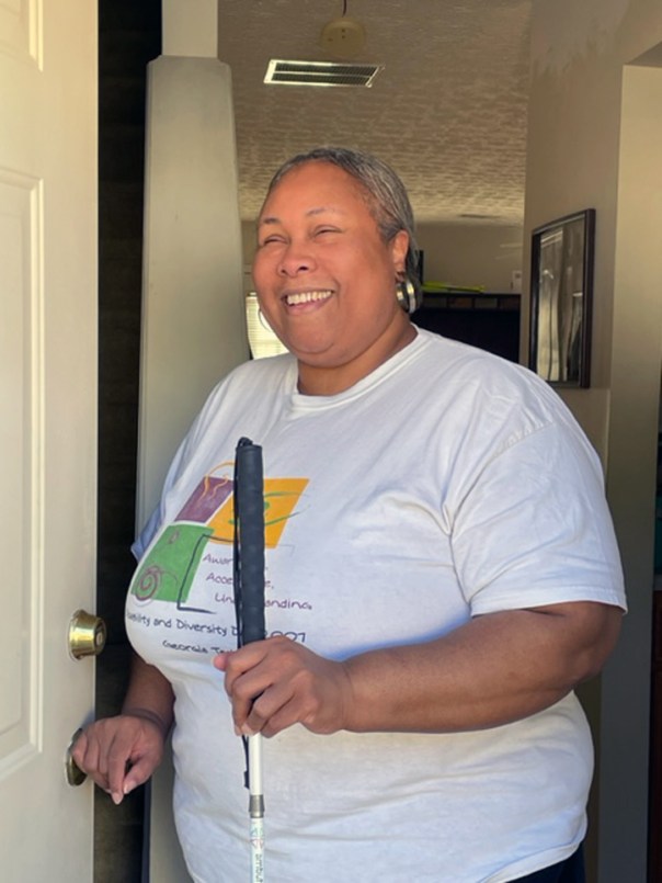 Empish is standing at her front door. She is holding her white cane and ready to leave out.