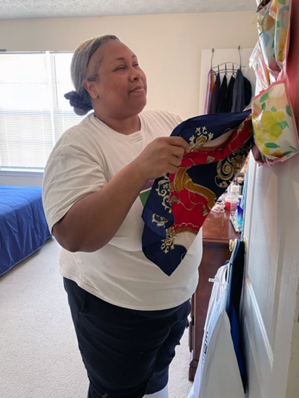 Empish is selecting a scarf from her hanging scarf organizer. It has several small pouches and some have braille labeling with a scarf inside.