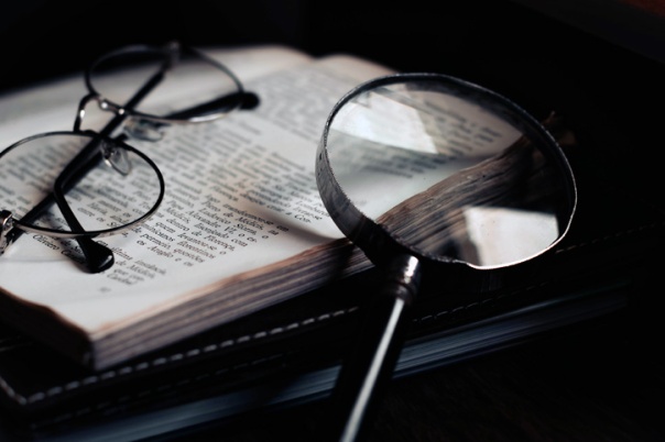 Sitting on a desk is a pair of glasses resting on an open book with a magnifier nearby.