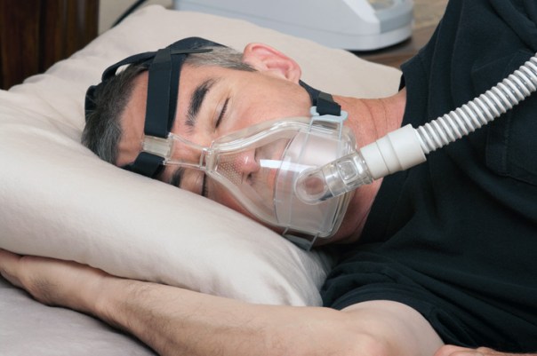 Man laying on his side in the bed with head on a pillow. His eyes are close. He is using a CPAP machine