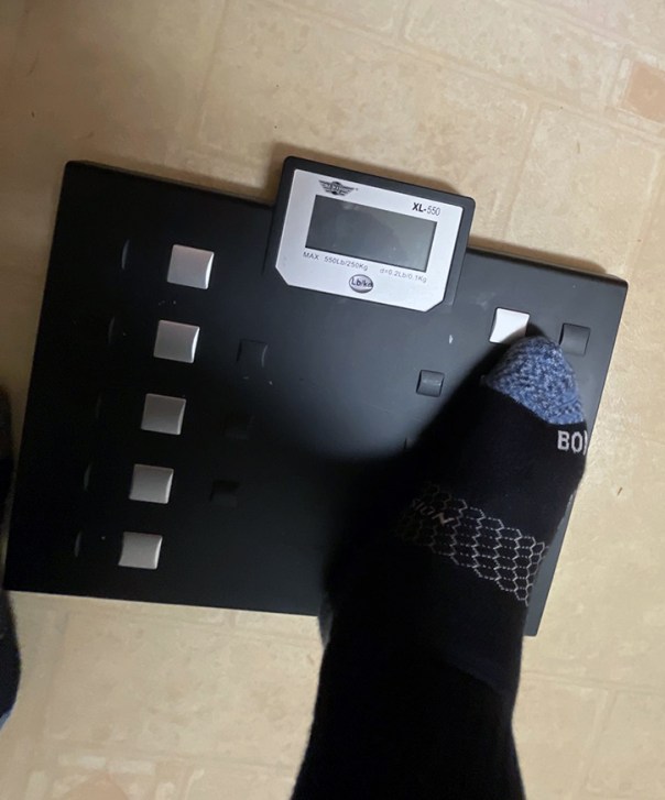 A bath scale sitting on the floor. It has one foot positioned on it.