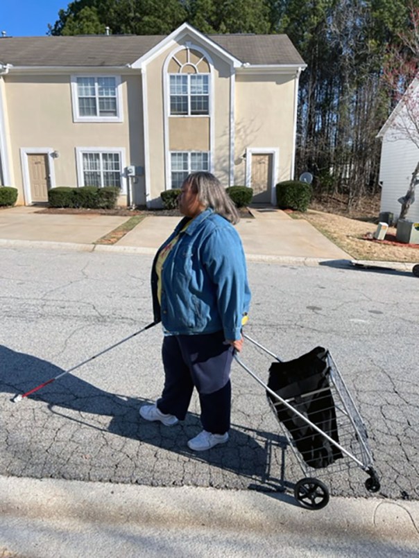 Empish is pulling her metal shopping cart in one hand while holding her white cane in another.