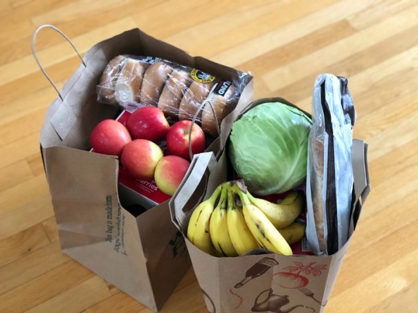 Two brown paper bags of groceries sitting on a hardwood floor. The bags contain apples, bag of bagels, a baguette and head of iceberg lettuce.