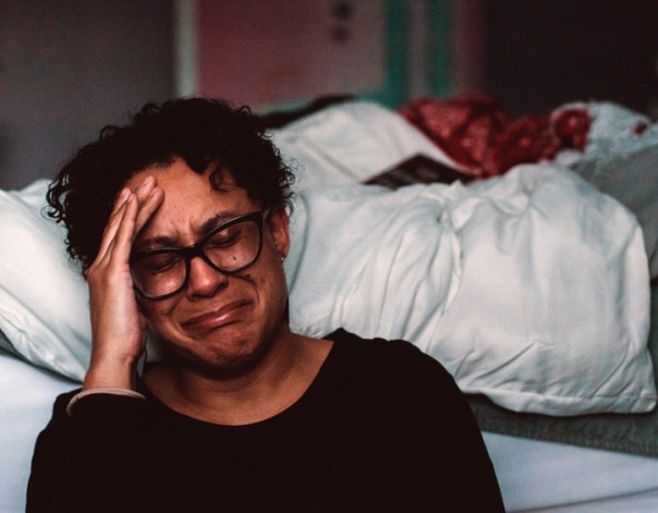 A woman crying. She is sitting on the floor beside her bed.
