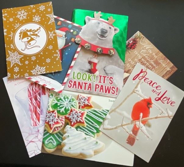 Several Christmas holiday cards are displayed on top of a table. One is a reindeer with a big nose and large antlers surrounded by white snowflakes. Another is a polar bear holding a red can and wearing a red and green scarf with bells. Another has Christmas cookies shaped like snowflakes with red and green frosting. Other cards have candy canes and other Christmas themes.